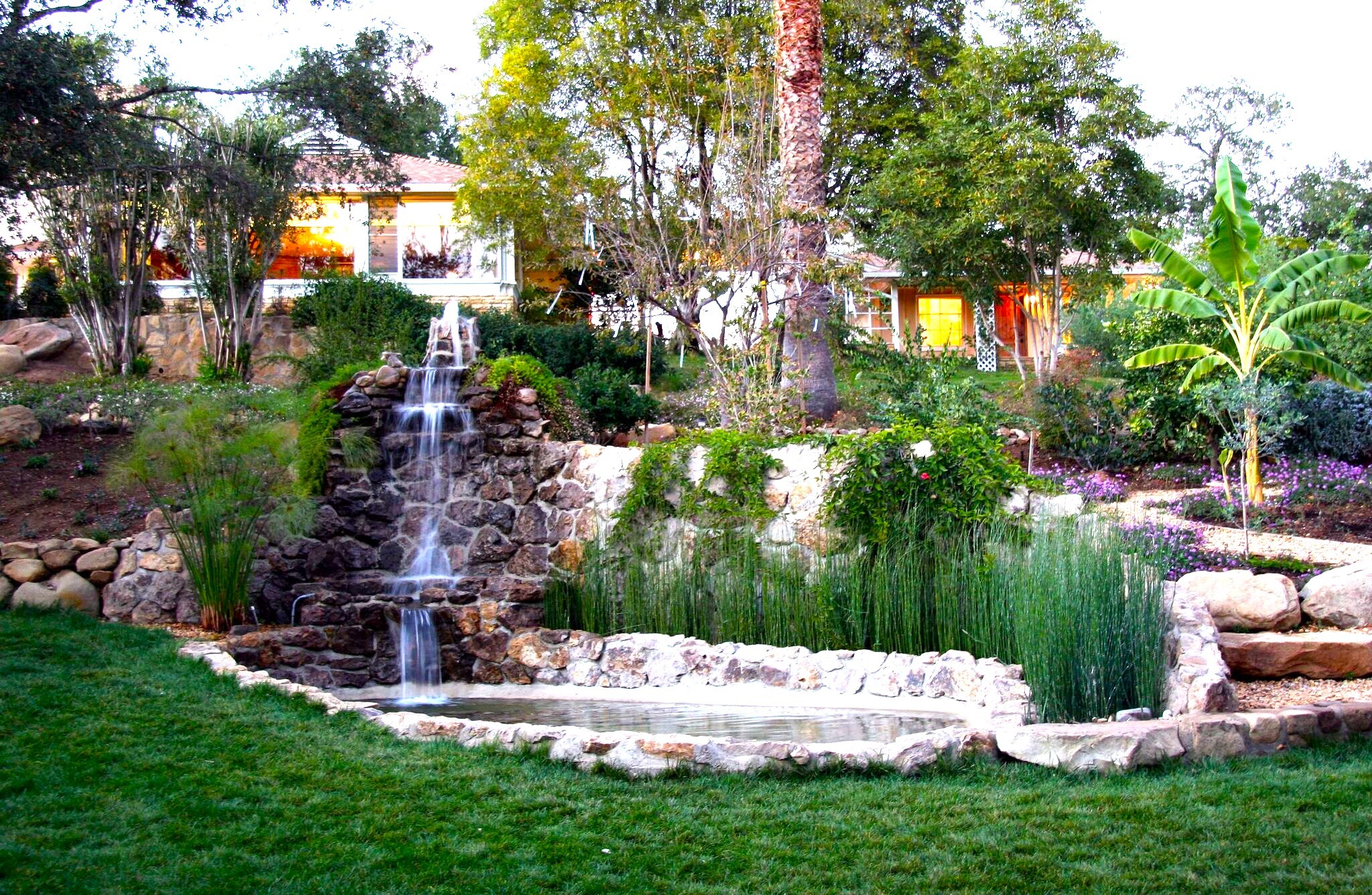 Garden with waterfall, trees, plants, and a grassy area, with houses in the background.