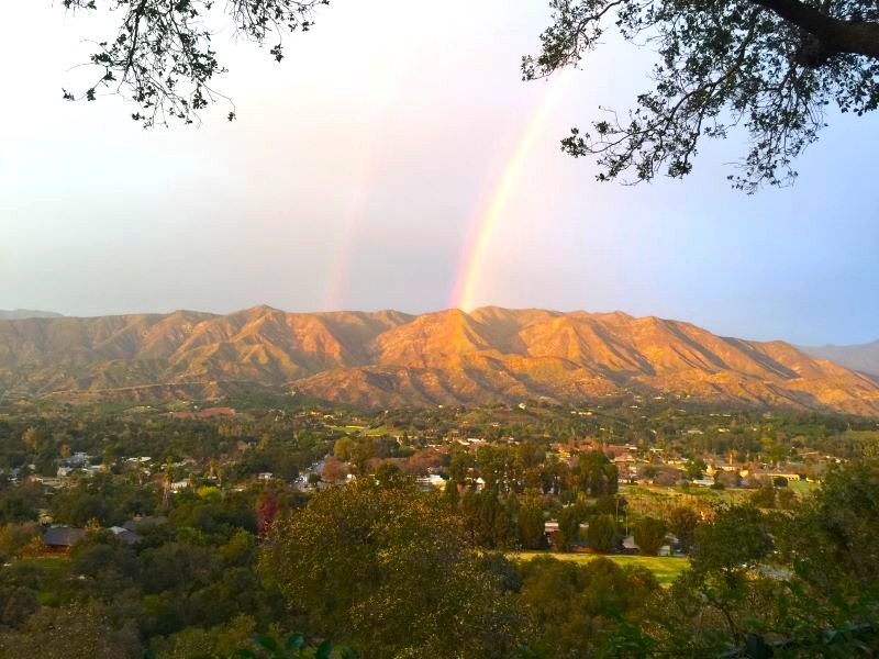 Landscape with mountains, rainbow, trees, and a city below, under a partly cloudy sky.