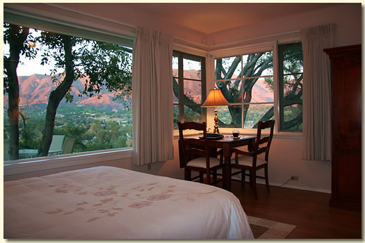 Bedroom with large windows showing trees and a sunset view, bed in foreground, small table with lamp and chairs near window.