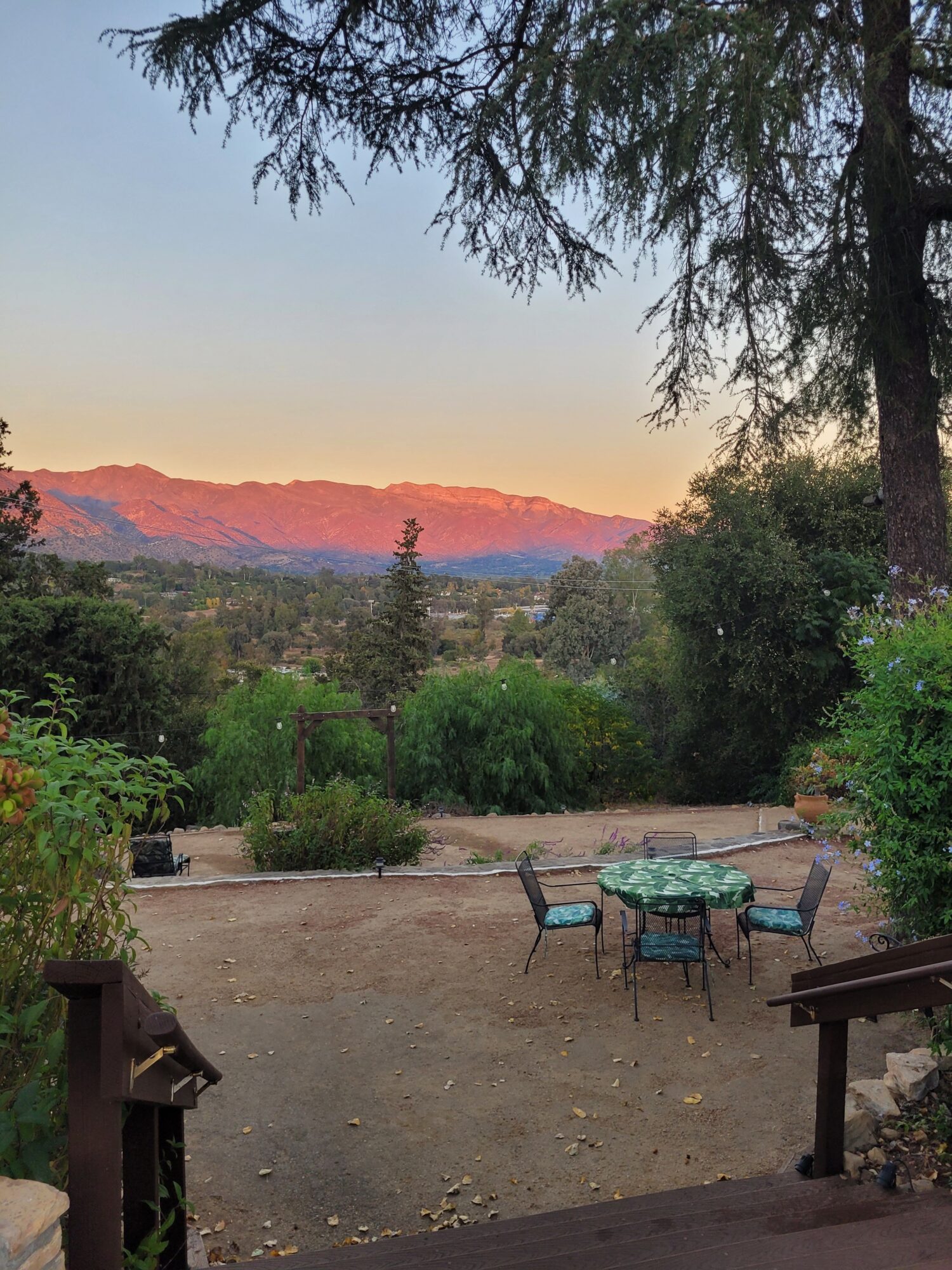 Sunset over mountains viewed from a patio with outdoor furniture and greenery.