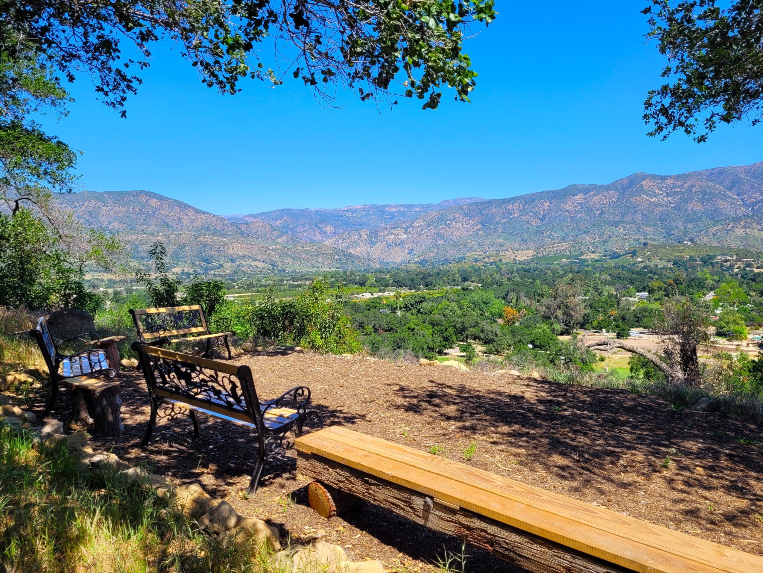 Three benches and a wooden platform on a dirt area overlooking a valley with mountains and trees under a blue sky.