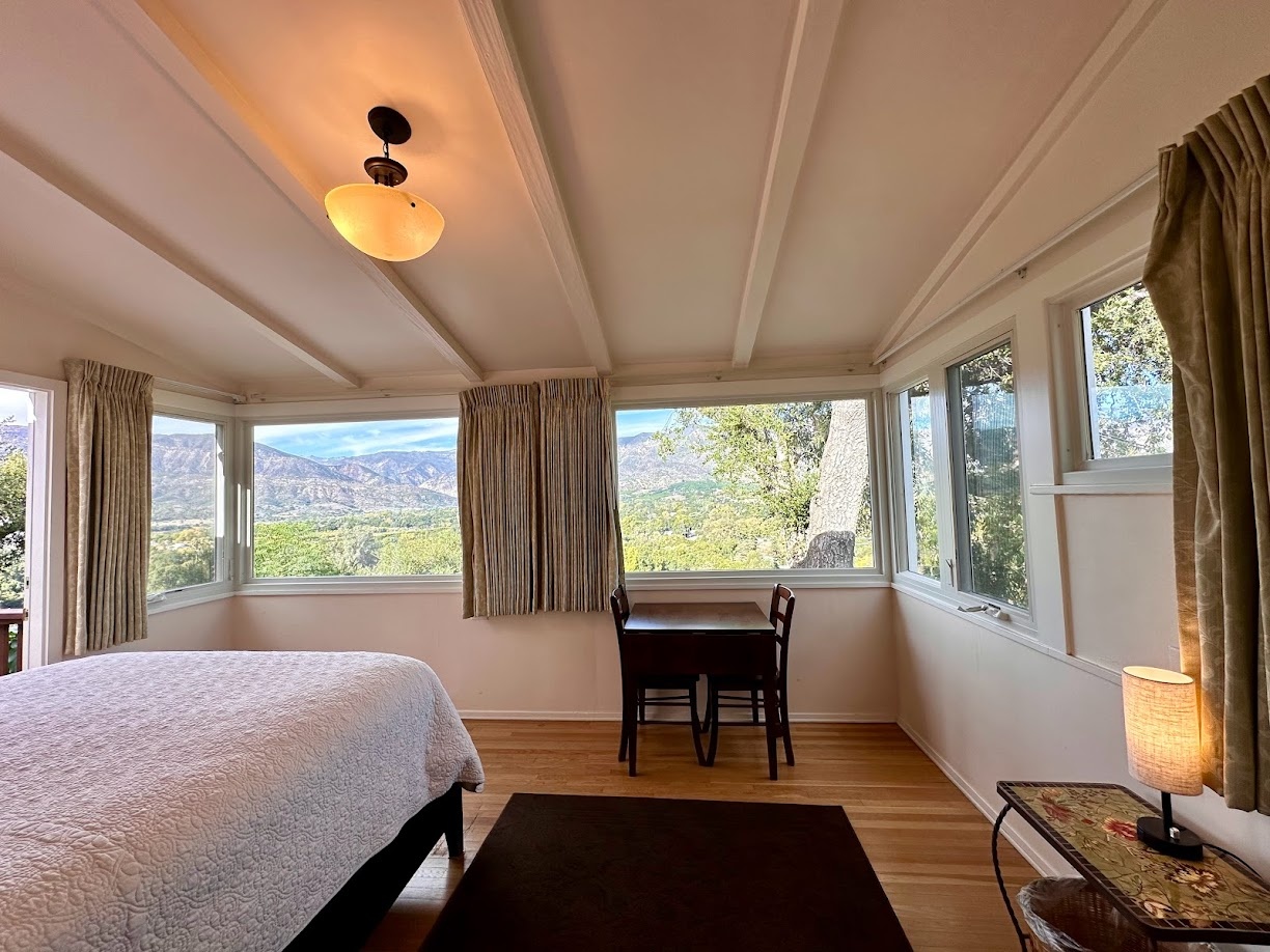 Room with large windows showing outdoor scenery, bed, desk, and a small table with a lamp, beige curtains, and wooden floor.