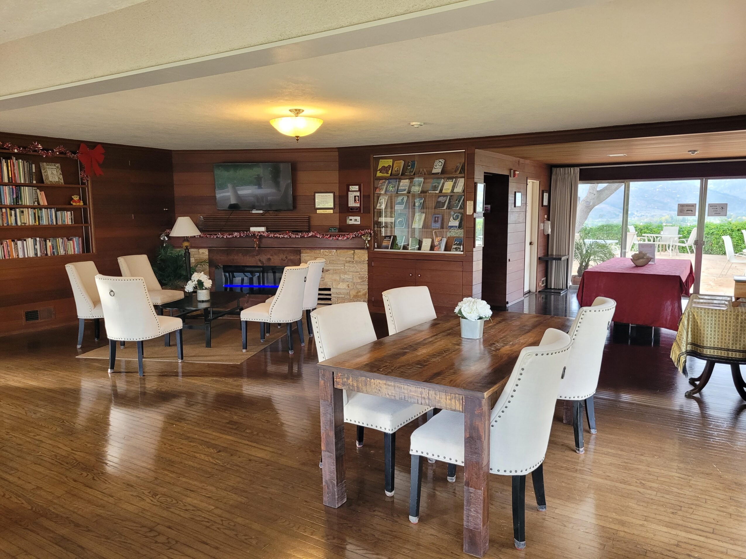 Room with wooden floors, a large dining table with six white chairs, a brick fireplace, a bookshelf, and a view outside through glass doors.