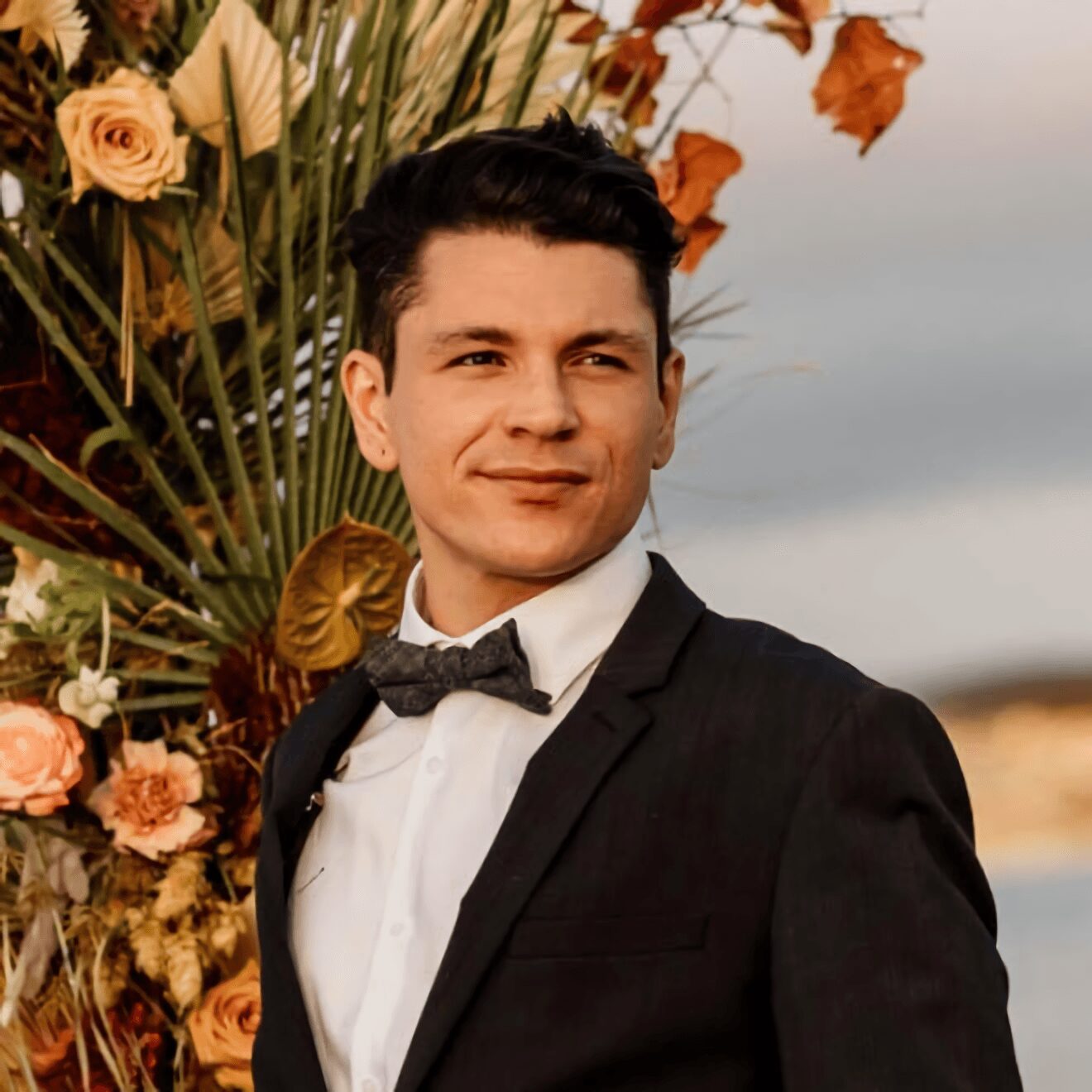 Young man in formal attire with a bow tie, standing outdoors near flowers and a body of water.