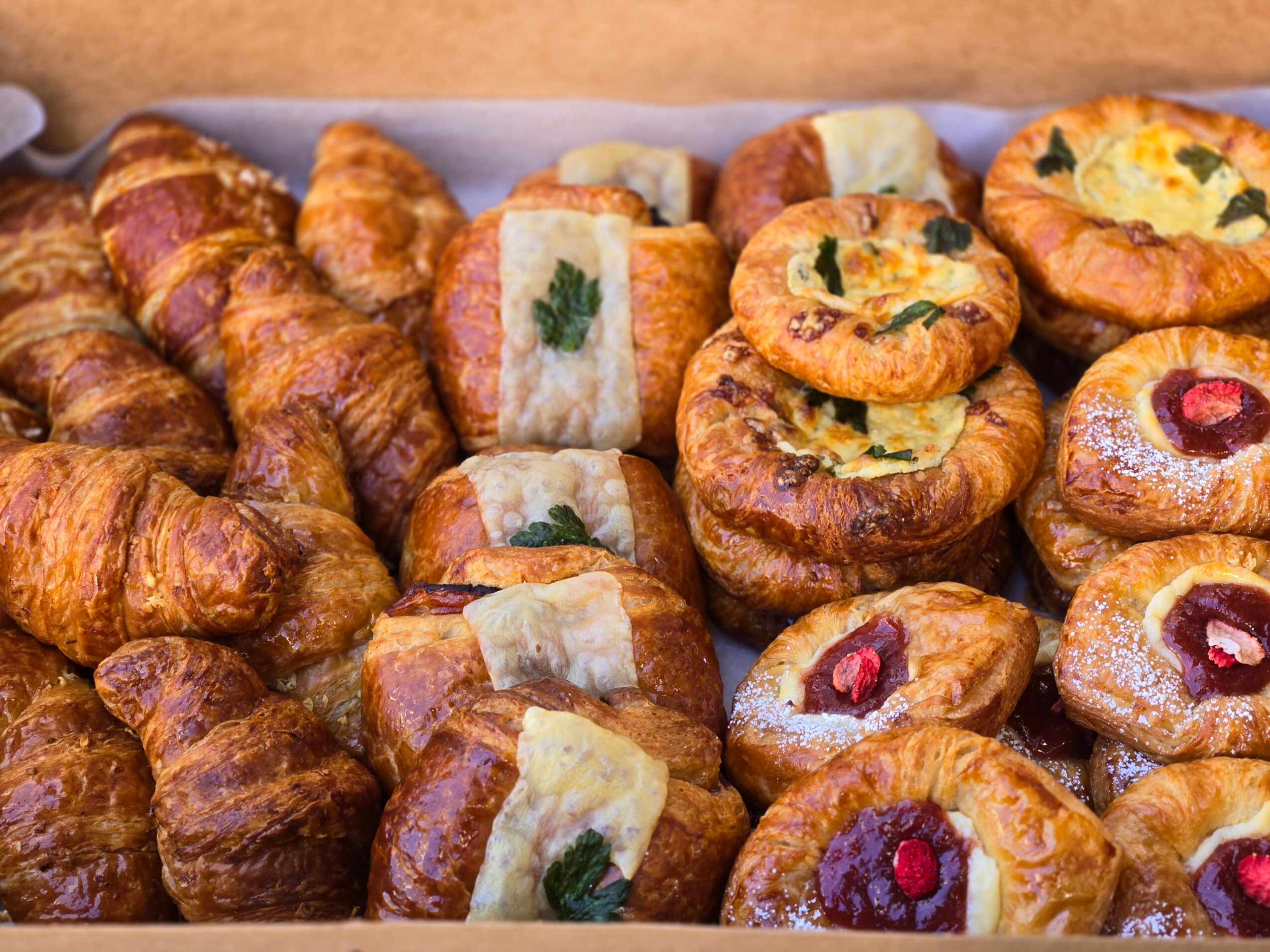 Assorted baked goods including croissants, small cakes with fruit toppings, and pastries with herbs and fillings, arranged on a tray.