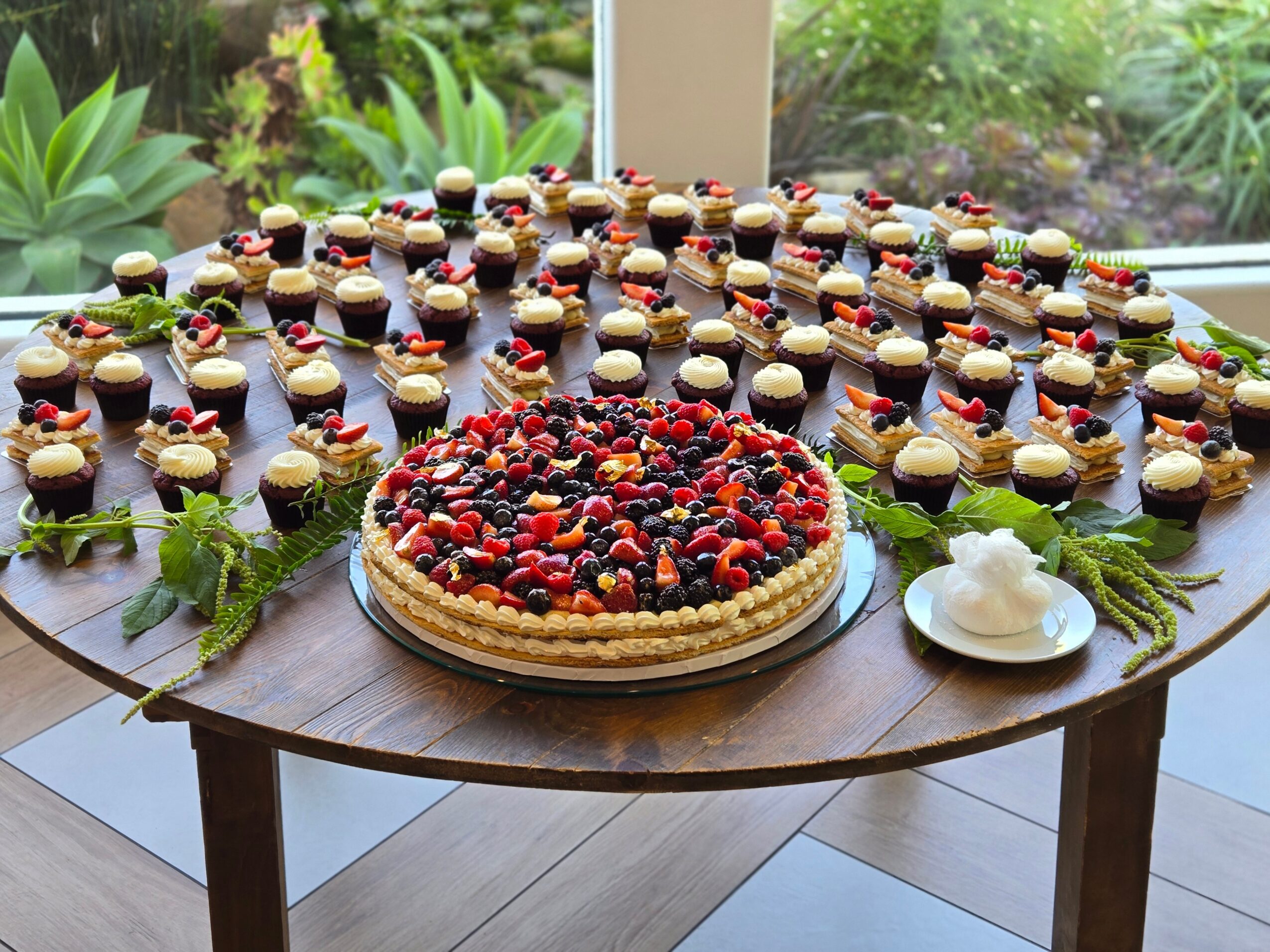 A round table with a large berry-topped cake, surrounded by small desserts, greenery, and a dollop of whipped cream, near a window.