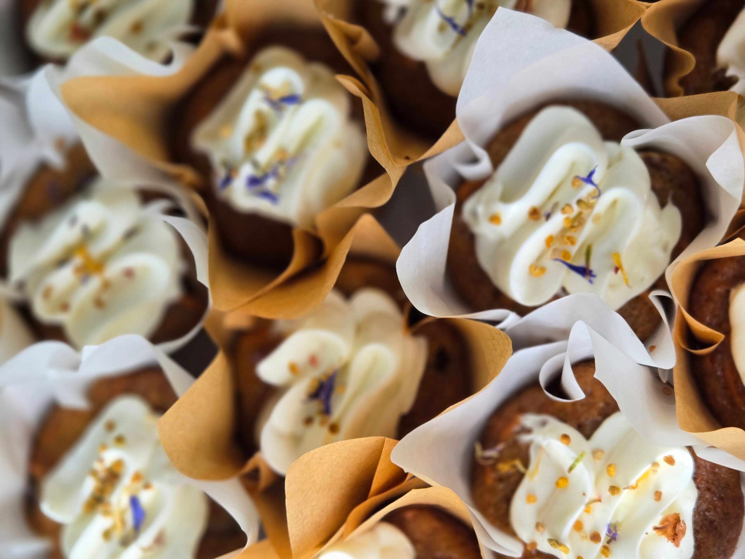 Multiple cupcakes with white frosting and colorful sprinkles, wrapped in brown and white paper, arranged closely together.