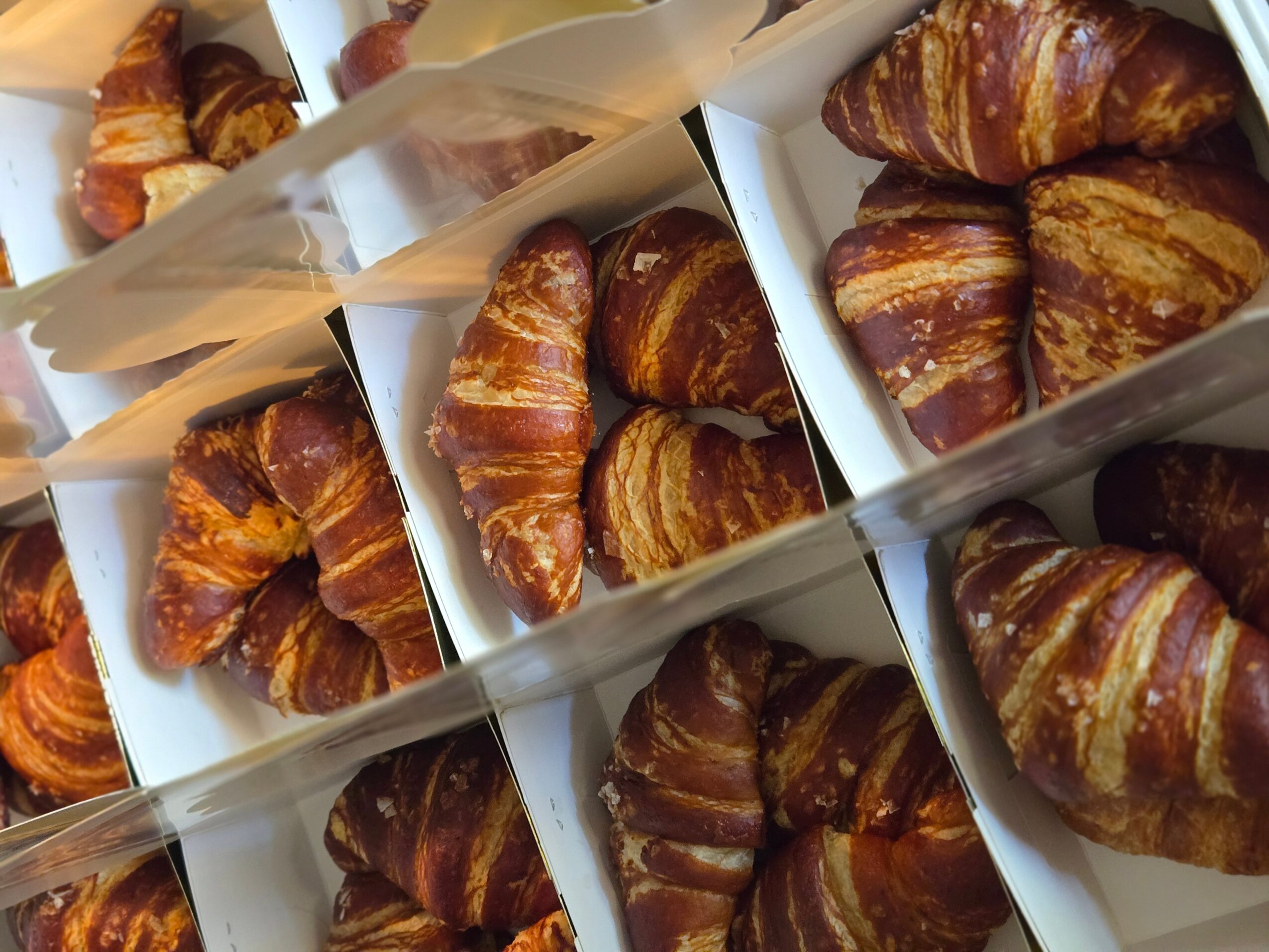 Multiple croissants in white boxes, stacked and arranged in rows, with golden-brown flaky layers visible.