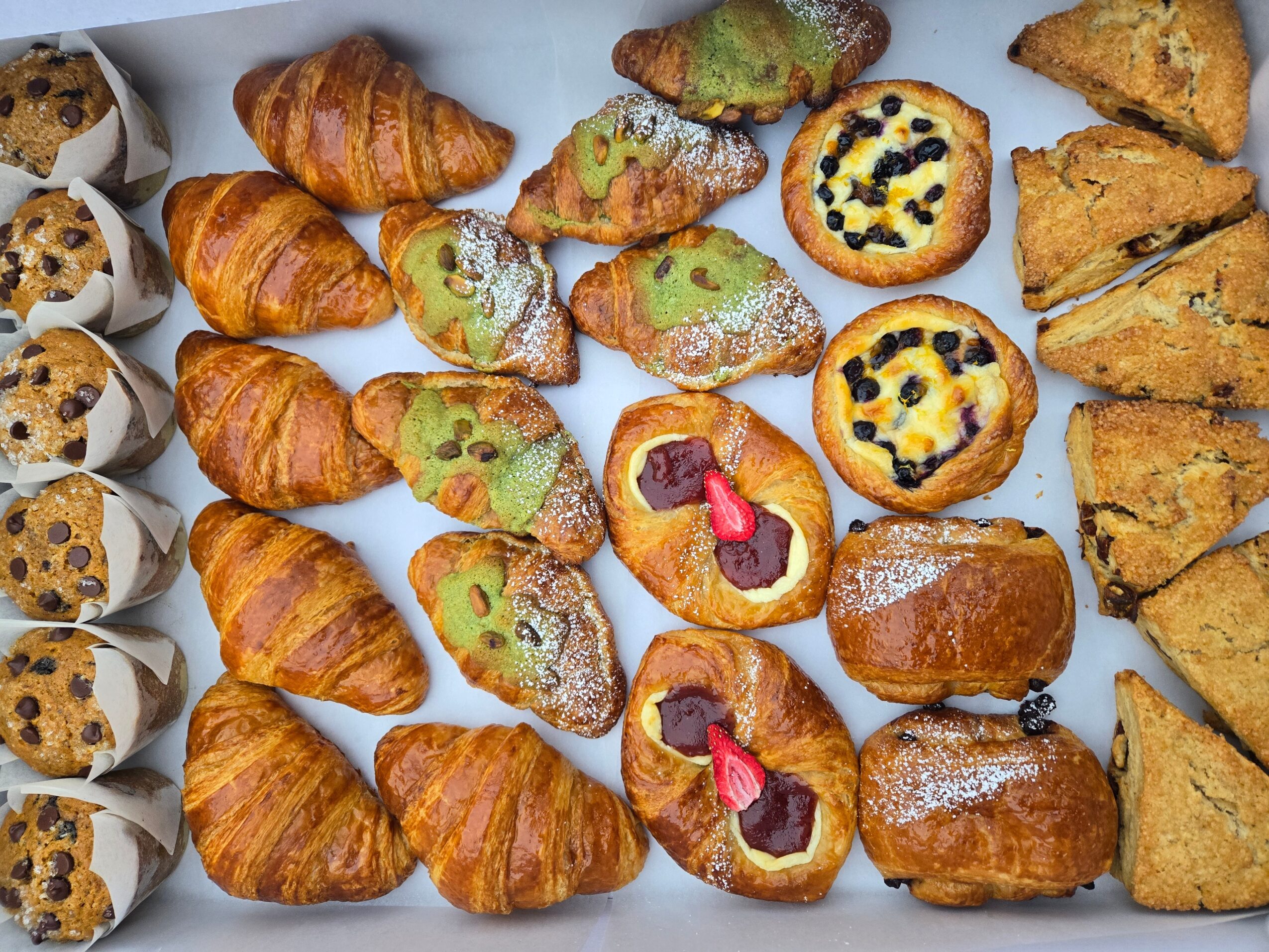Assorted baked goods including croissants, cookies, muffins, and scones arranged on a white surface.