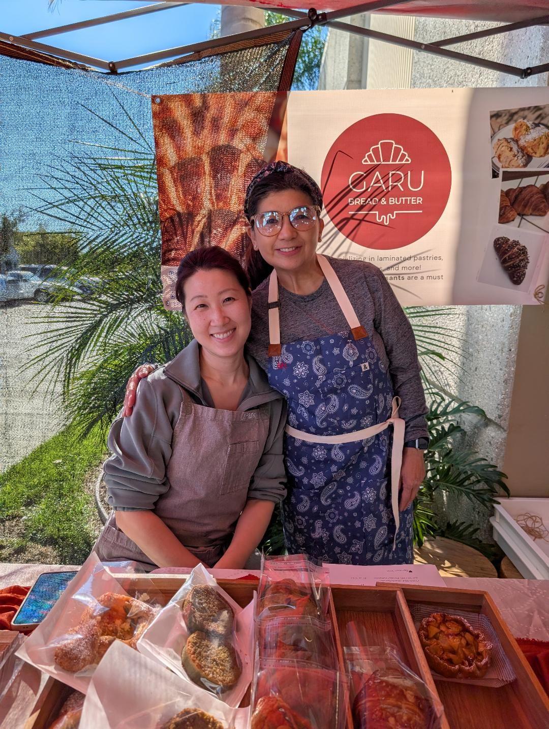 Two women wearing aprons stand behind a food stall with various baked goods, smiling at the camera, outdoors with plants and a banner in the background.