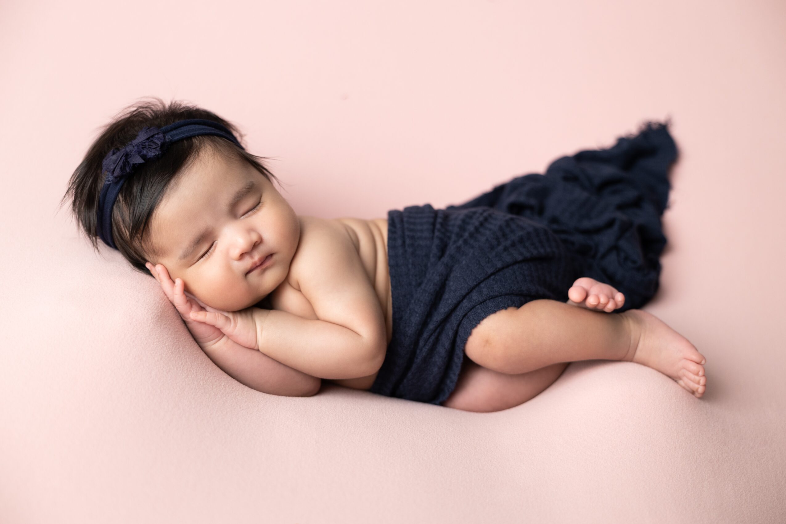 Baby sleeping on pink surface, wearing a dark headband and navy wrap, with a dark ruffled blanket behind.