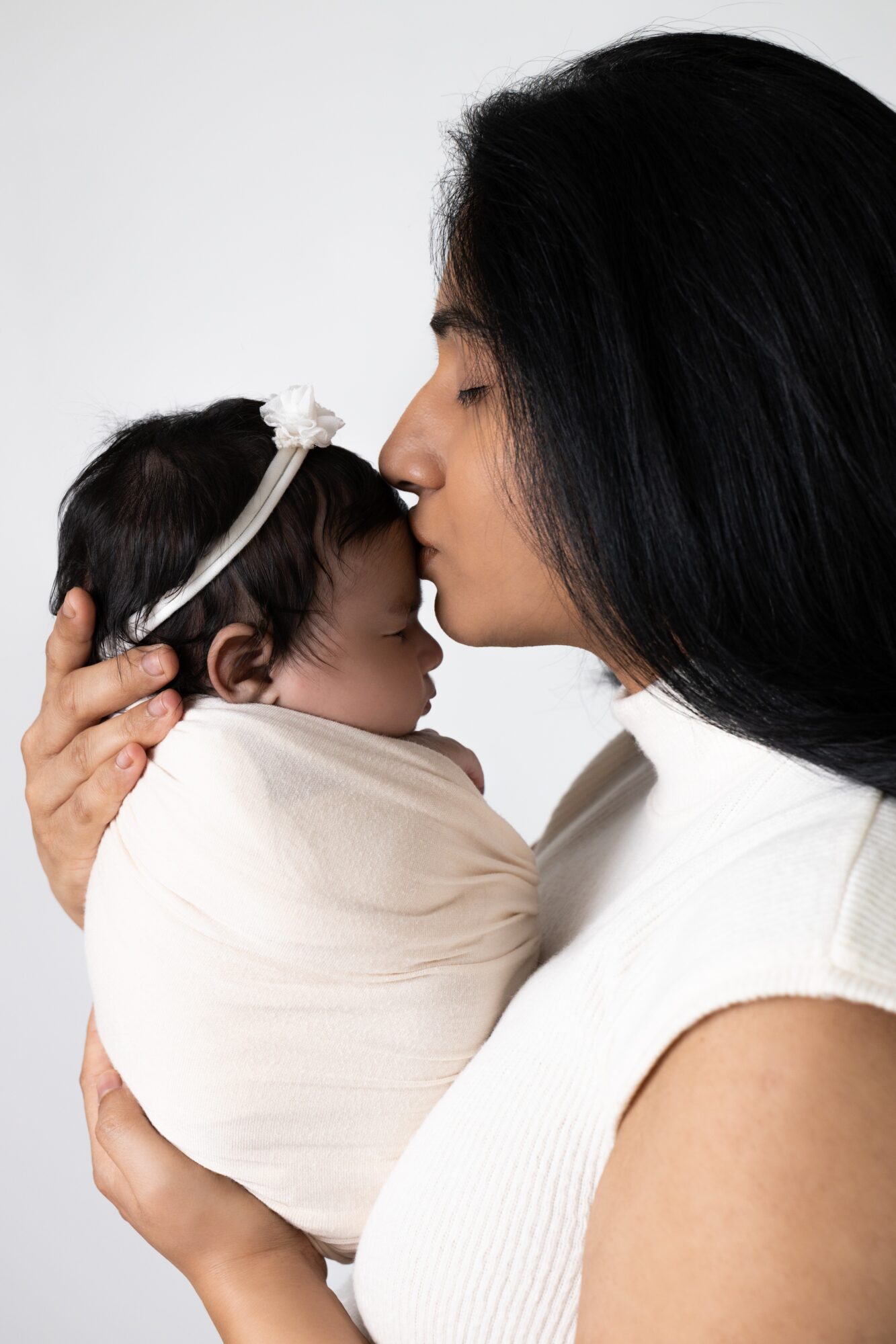 Woman holding a sleeping baby wrapped in a blanket, gently touching her forehead to the baby's head.