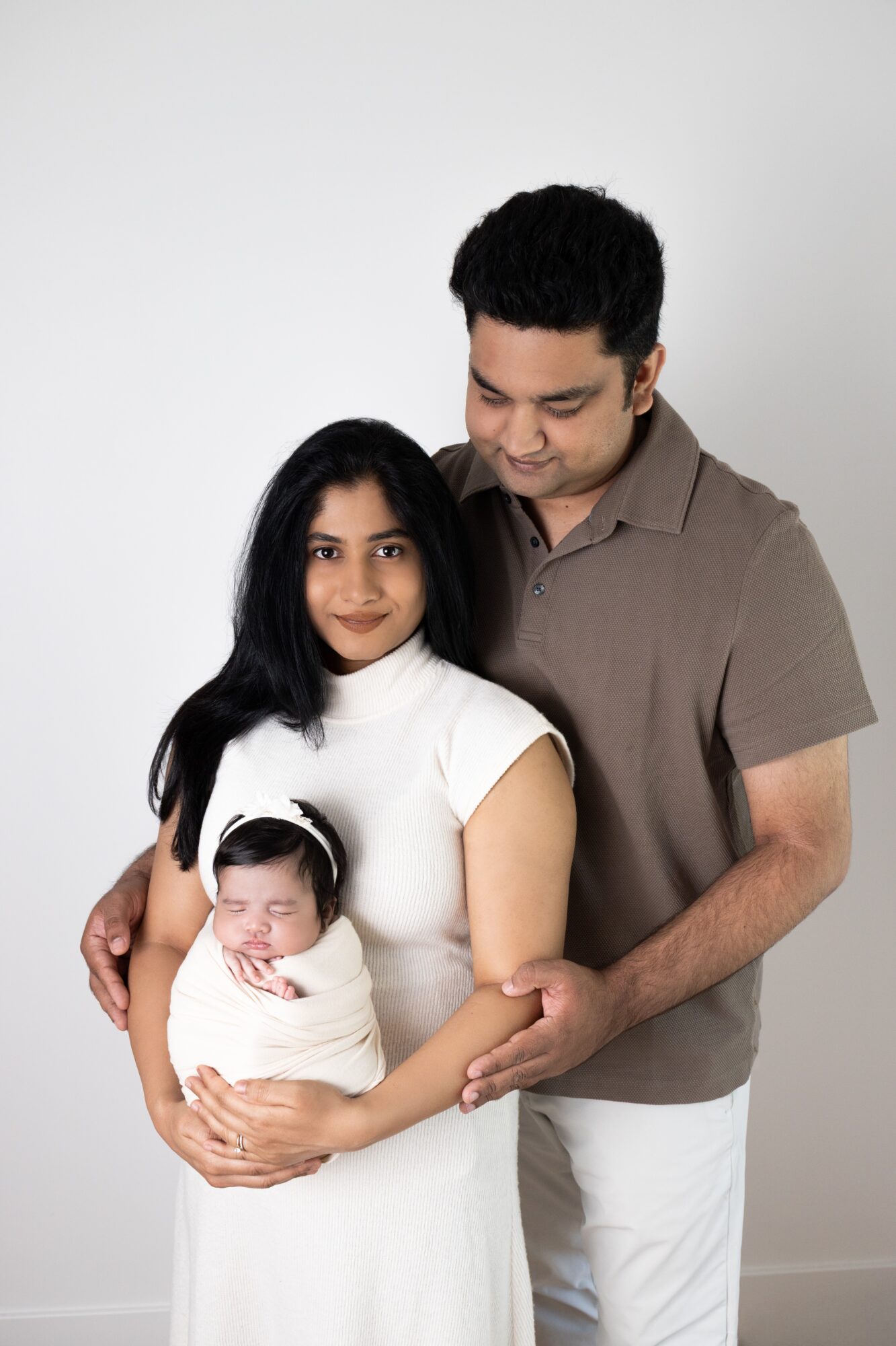 Family of three with a woman holding a newborn, man standing beside her, all smiling, against a plain white background.