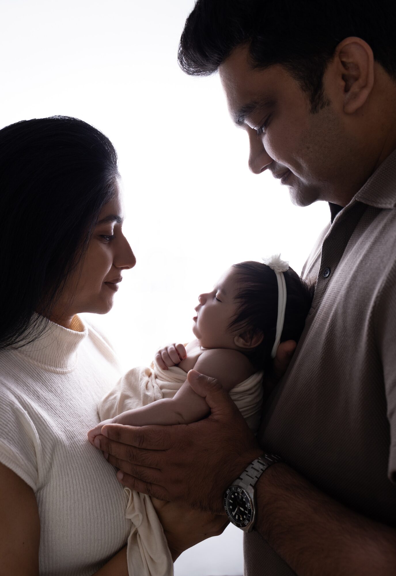 A man and woman look at a baby they are holding, with a white background.