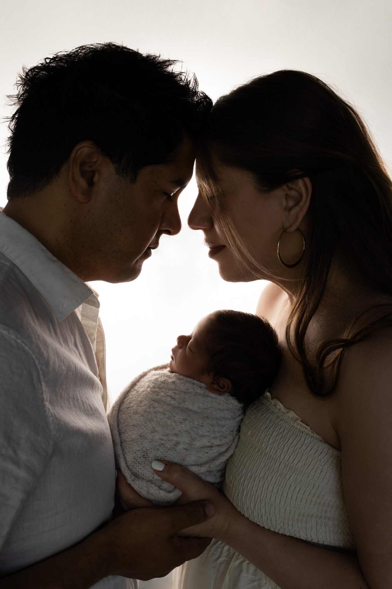 Silhouette of a family with a newborn, faces close together, backlit background, woman holding baby, man touching baby, intimate scene.