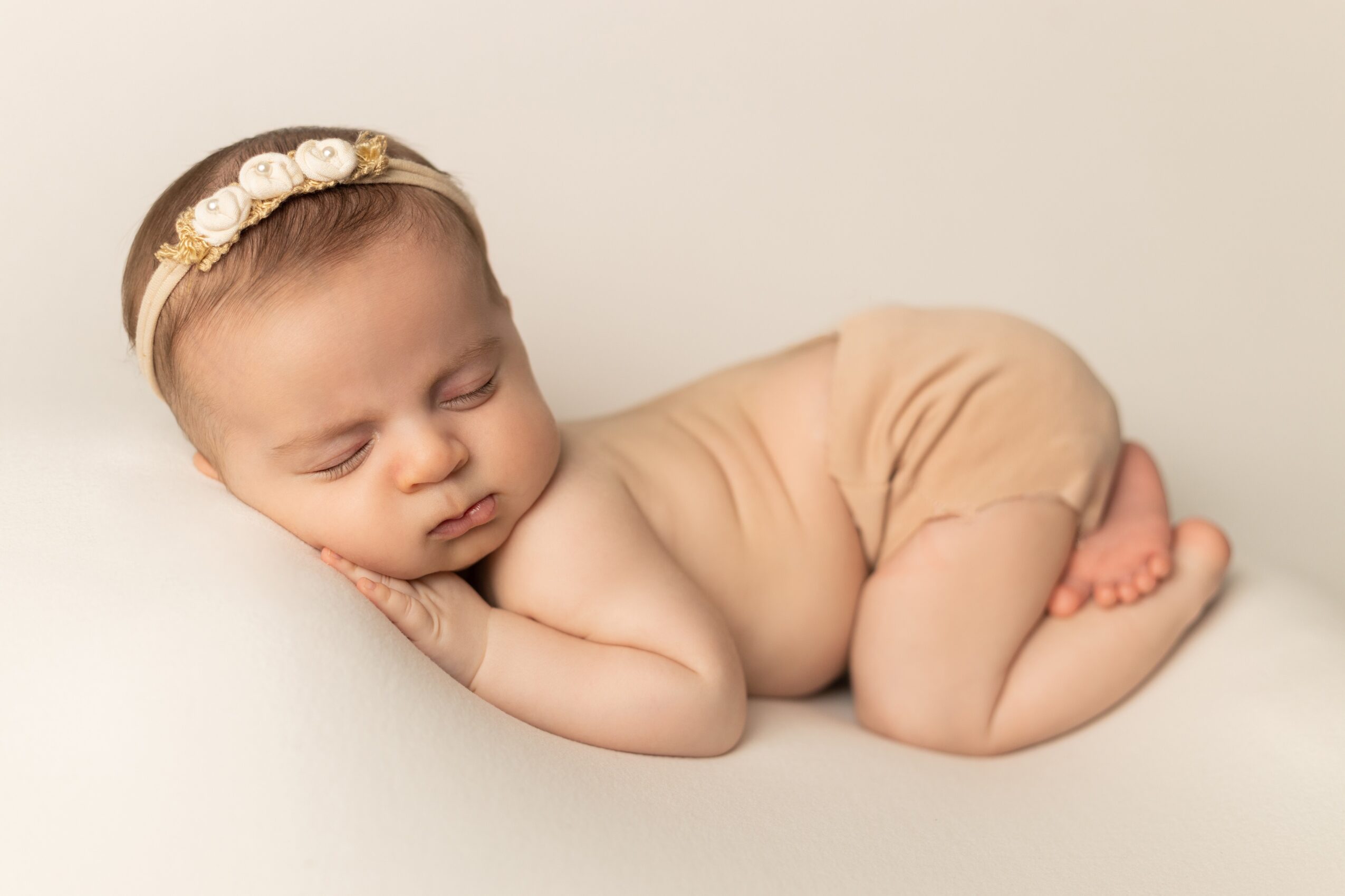 Baby girl sleeping on her side with a floral headband, lying on a soft surface, wearing beige shorts.