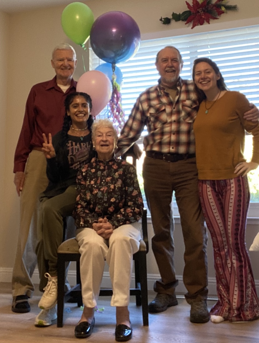 Group of five people, four standing and one sitting, smiling indoors with balloons and a window in the background.
