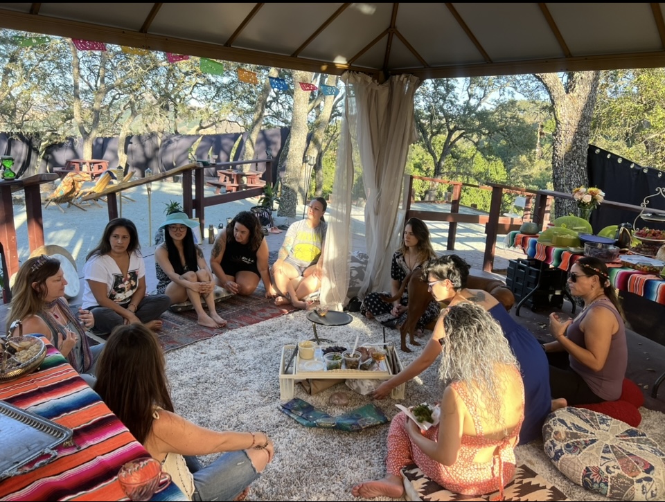 Group of people sitting in a circle under a canopy outdoors, with trees and a wooden deck in the background.