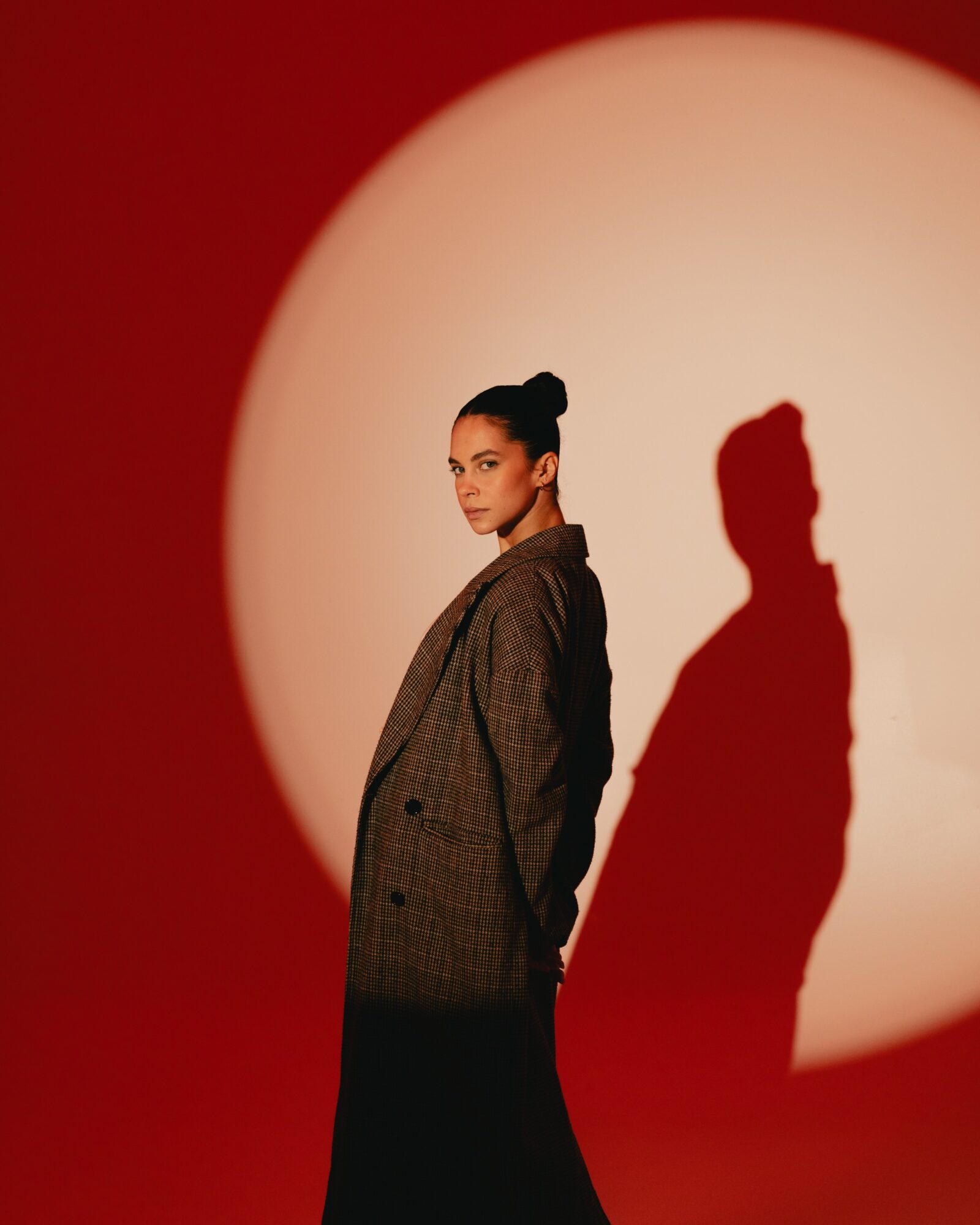 Woman with dark hair in a bun wearing a blazer, standing against a red background with a large white circle and shadow.