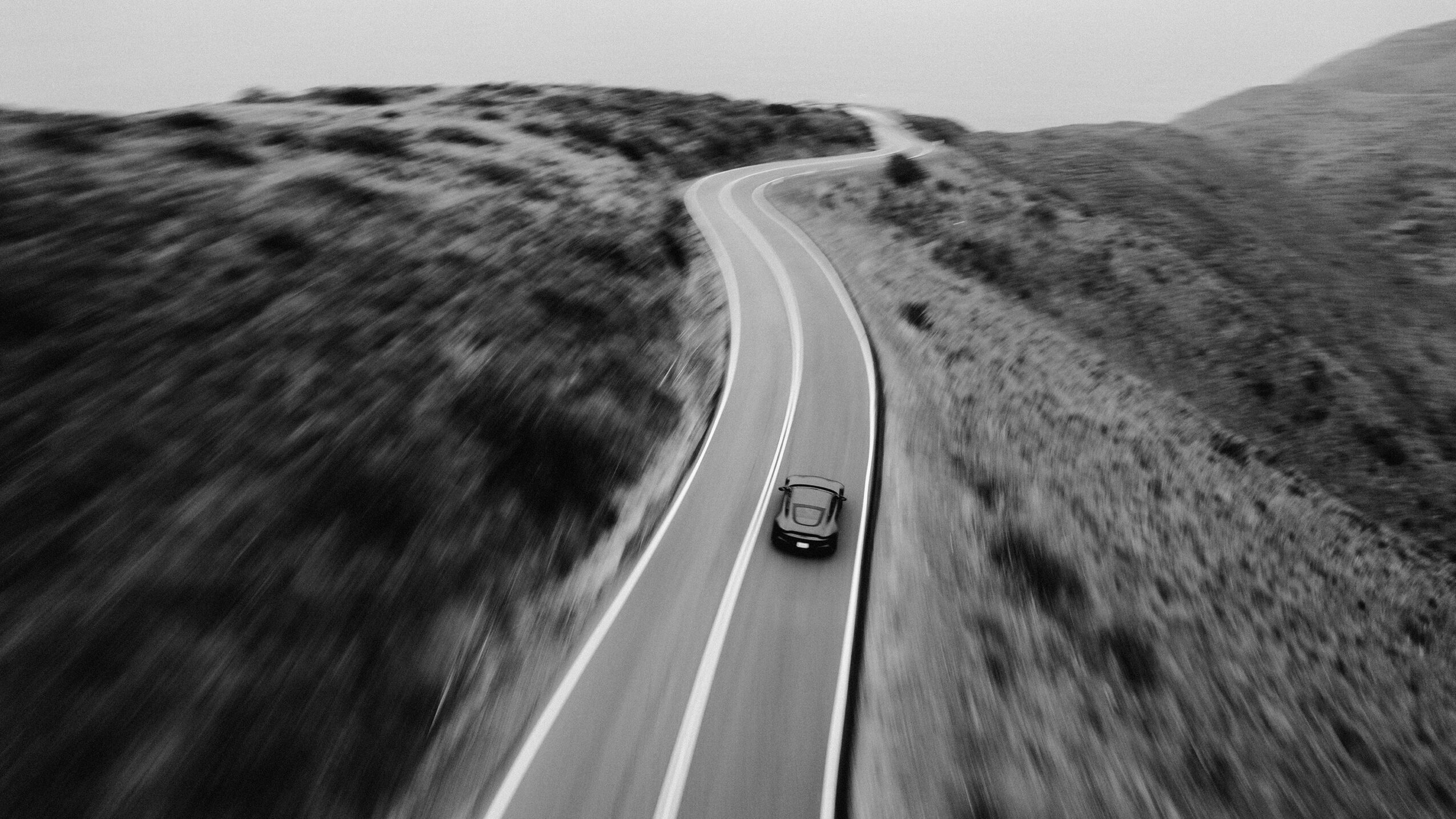 A car driving on a winding road through hilly terrain, black and white image, viewed from above.
