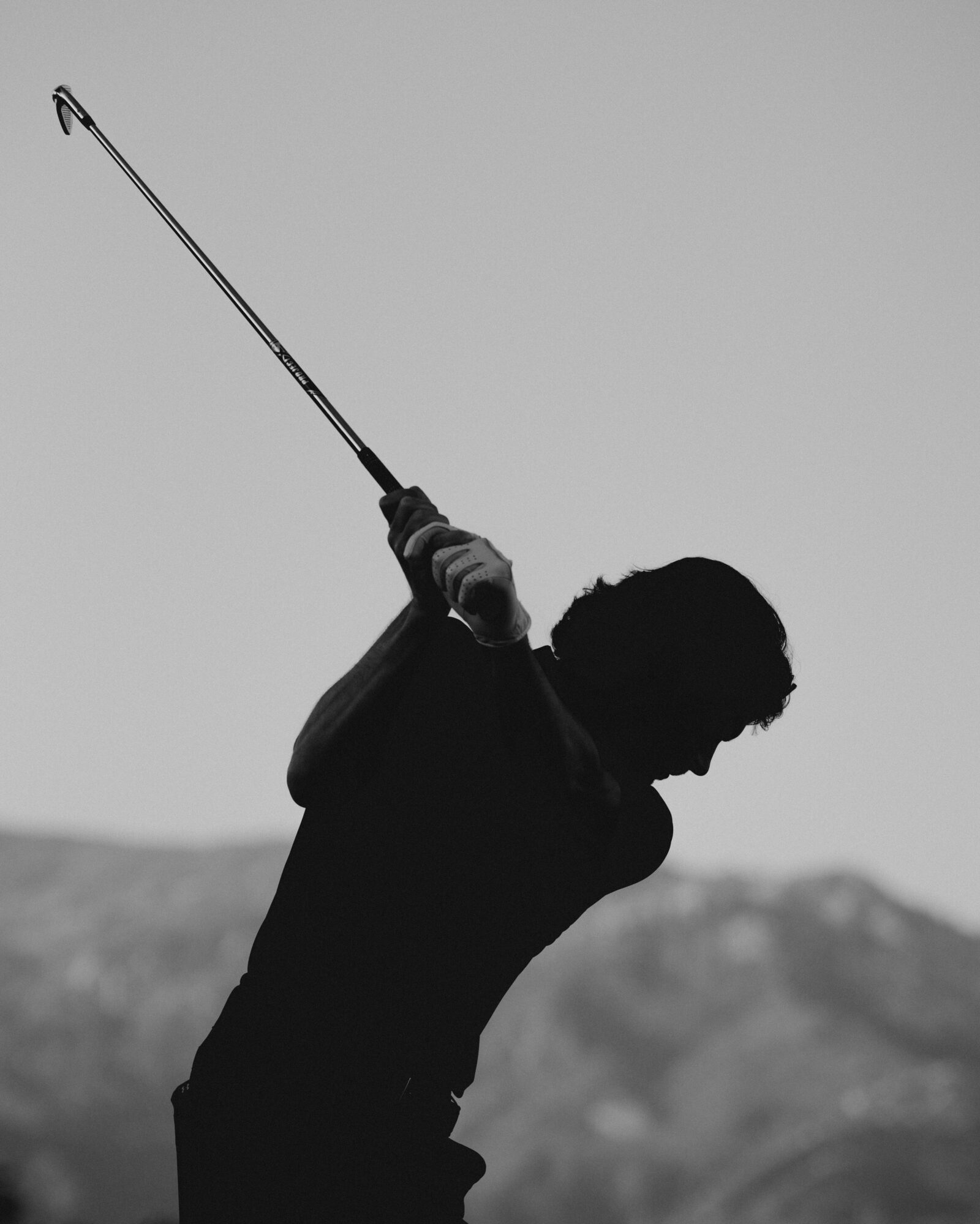 Silhouette of a person swinging a golf club against a mountain landscape, sky in the background.