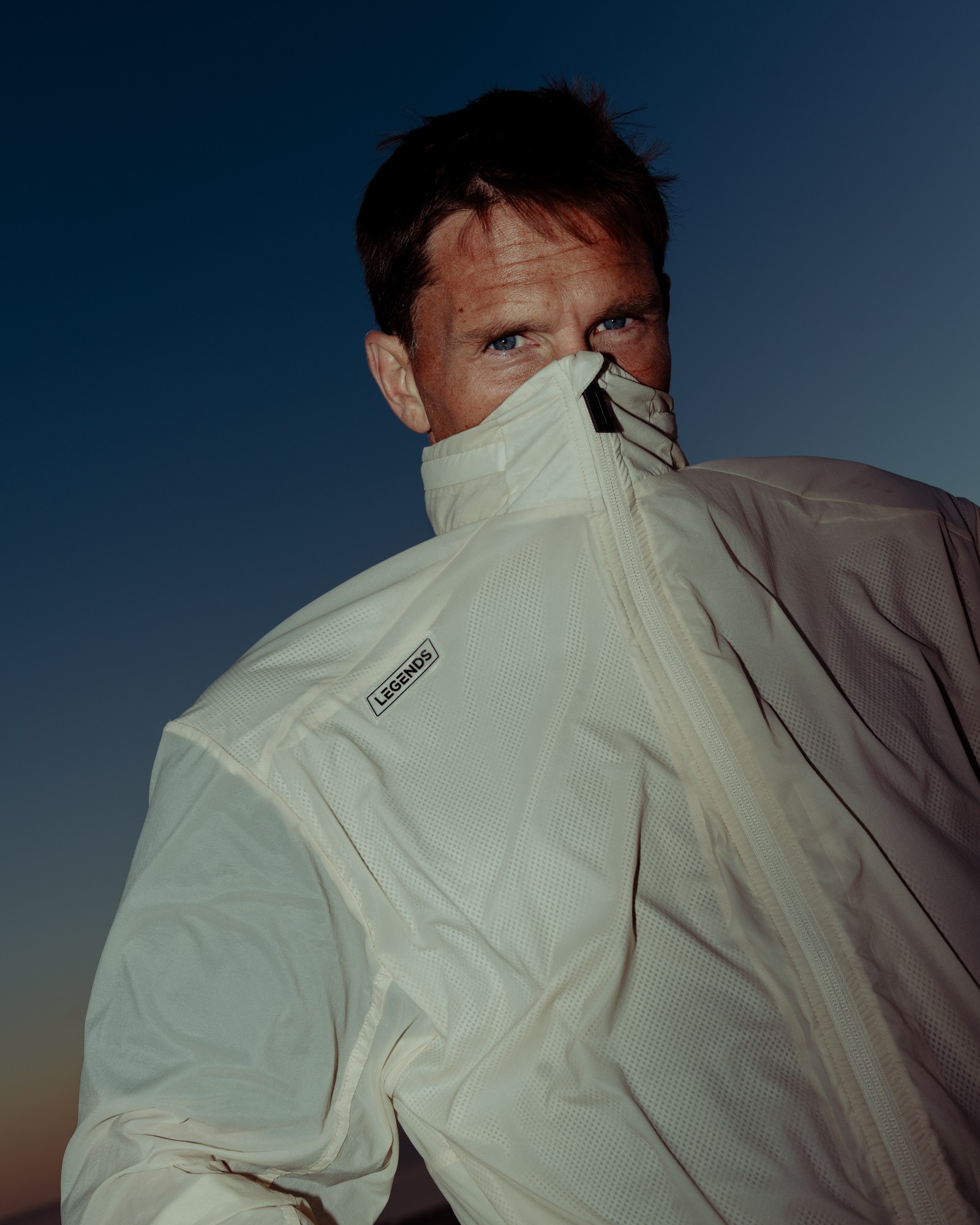 Person wearing a white jacket with the collar zipped up, partially covering their face, against a dark sky background.