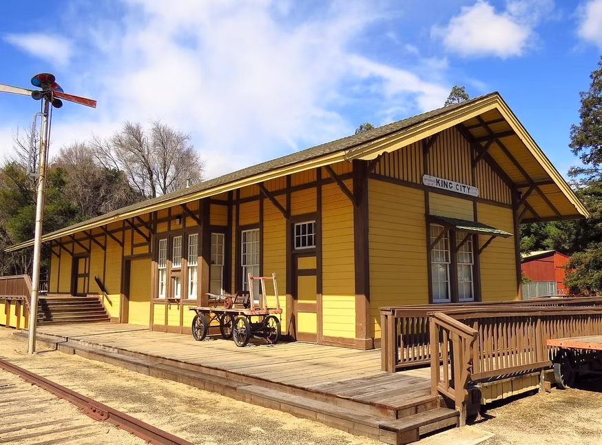 A yellow train station building with a gabled roof, windows, and a platform with a cart, under a blue sky.