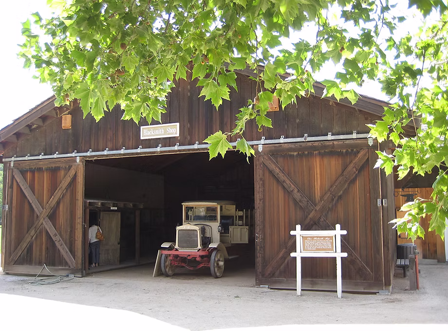 Barn with open doors, a small vehicle parked inside, and a white signboard outside, partially obscured by green leaves.