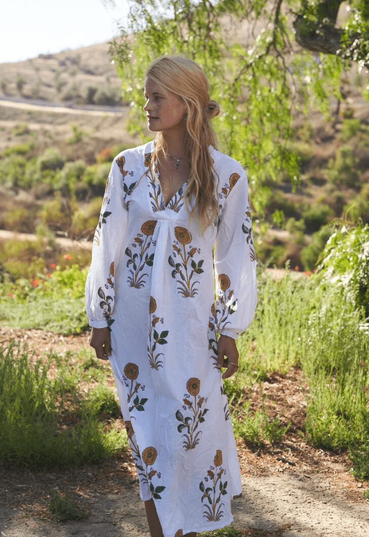 Woman with long blonde hair wearing a white dress with floral patterns outdoors, trees and hills in background.