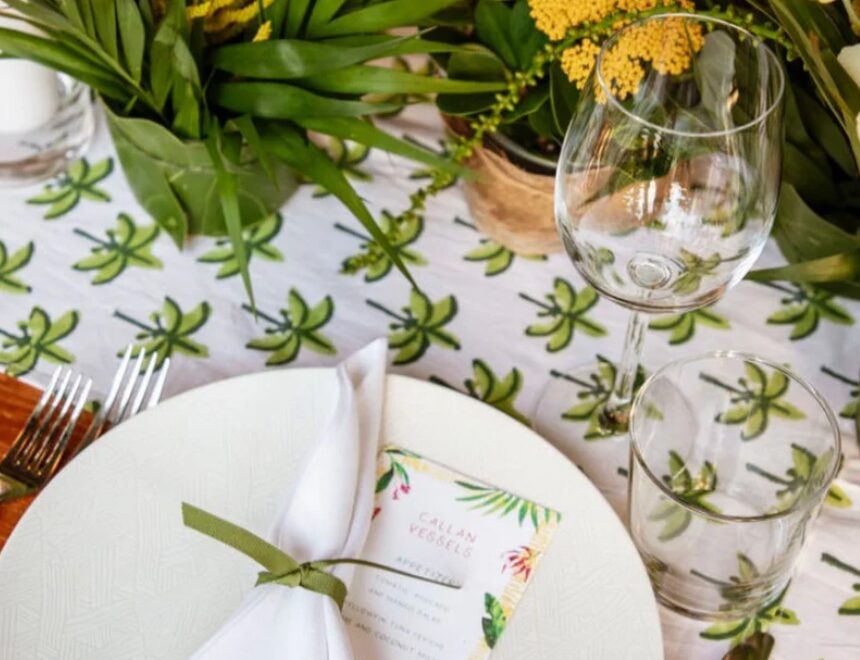 Table setting with a white plate, napkin, glass, and greenery, with a floral menu and plants in the background.
