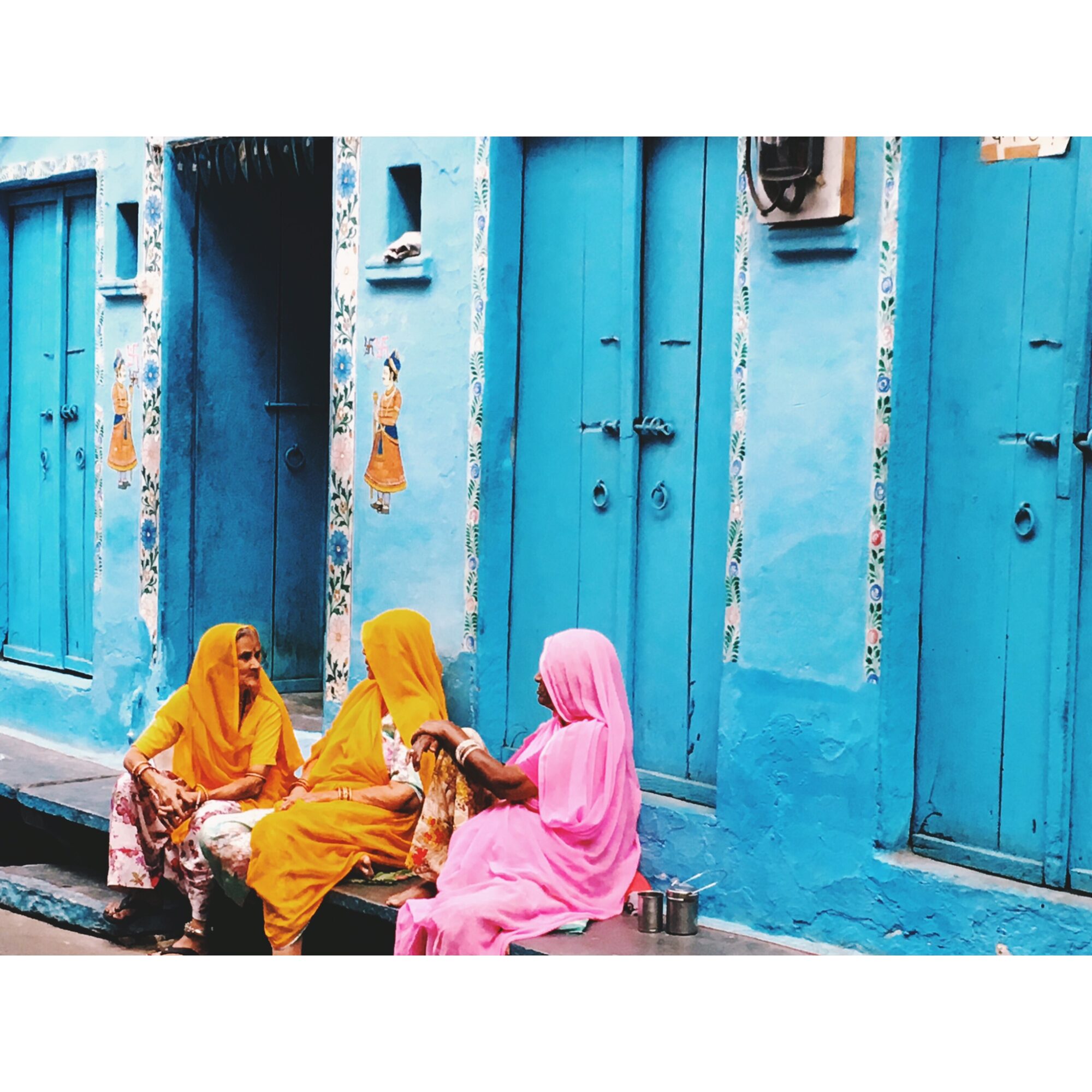 Three women in colorful traditional clothing sit against a bright blue wall with doors, engaged in conversation.