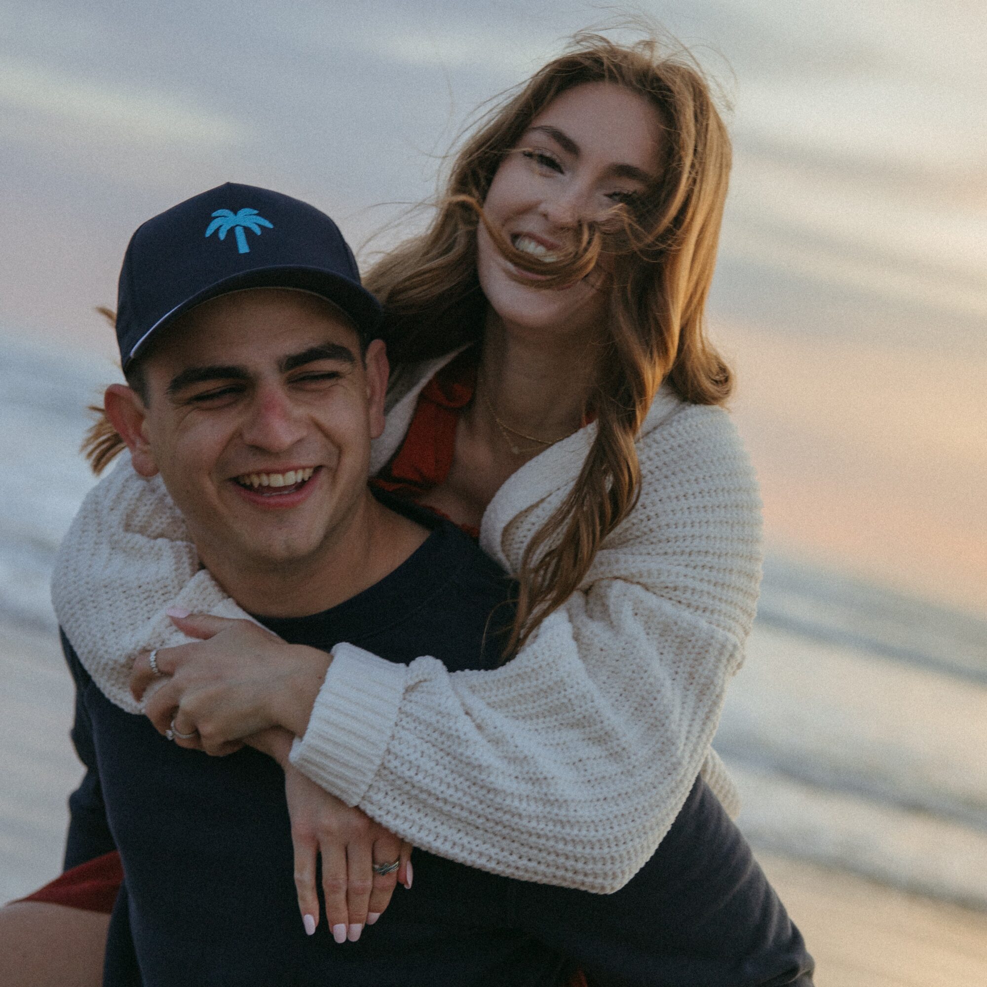 A smiling young man and woman at the beach during sunset, with the woman hugging the man from behind.