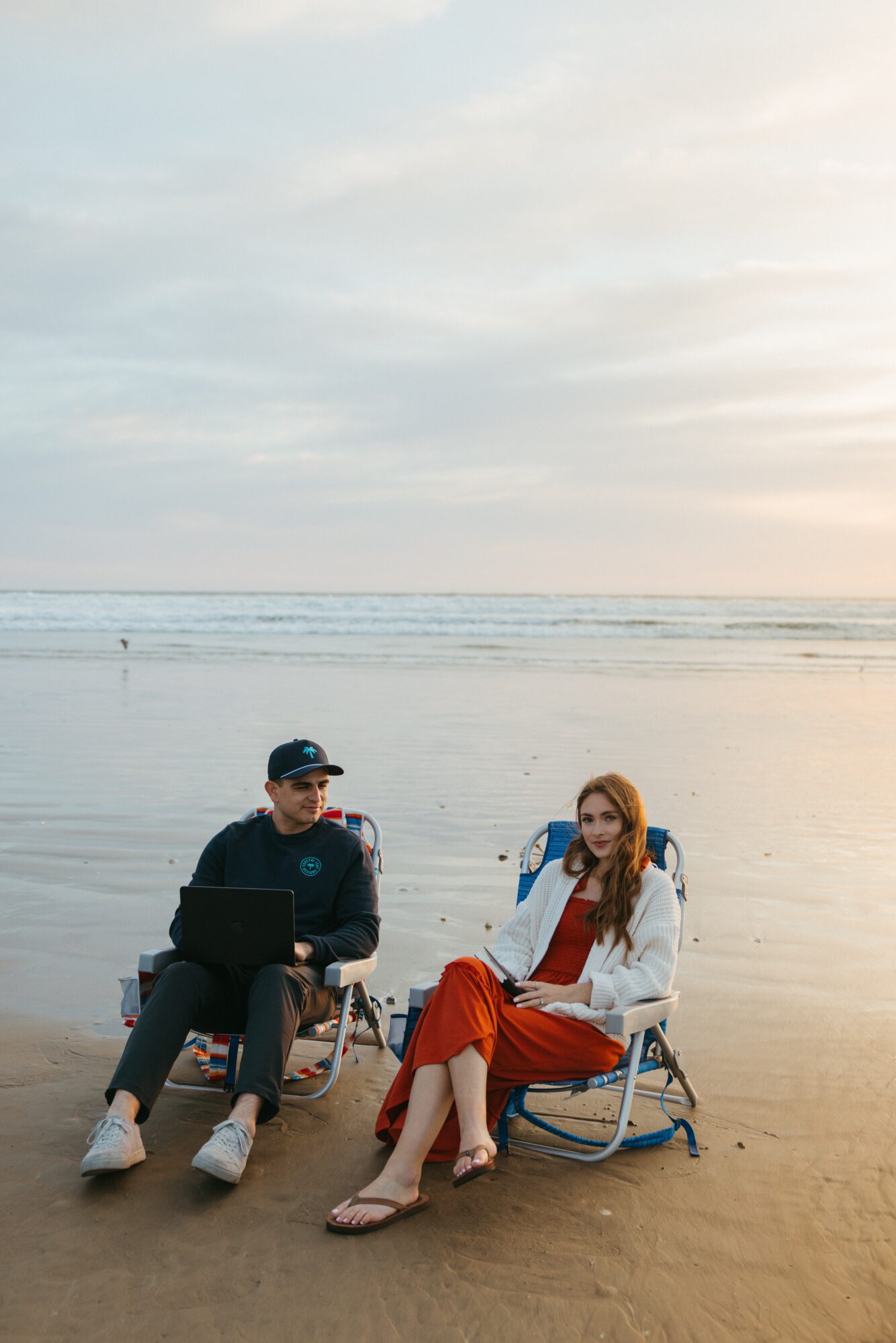 Two people sit on chairs on a beach with the ocean and sky in the background, one using a laptop and the other talking.