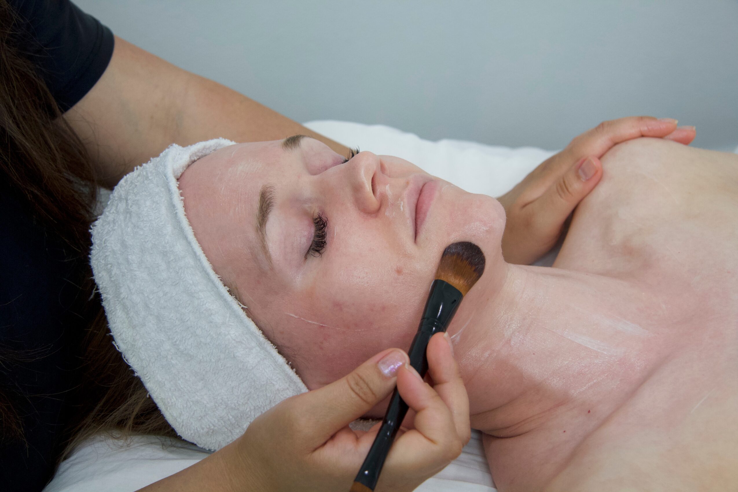 Woman receiving facial treatment with a brush on her face, wearing a towel wrapped around her head, lying down.