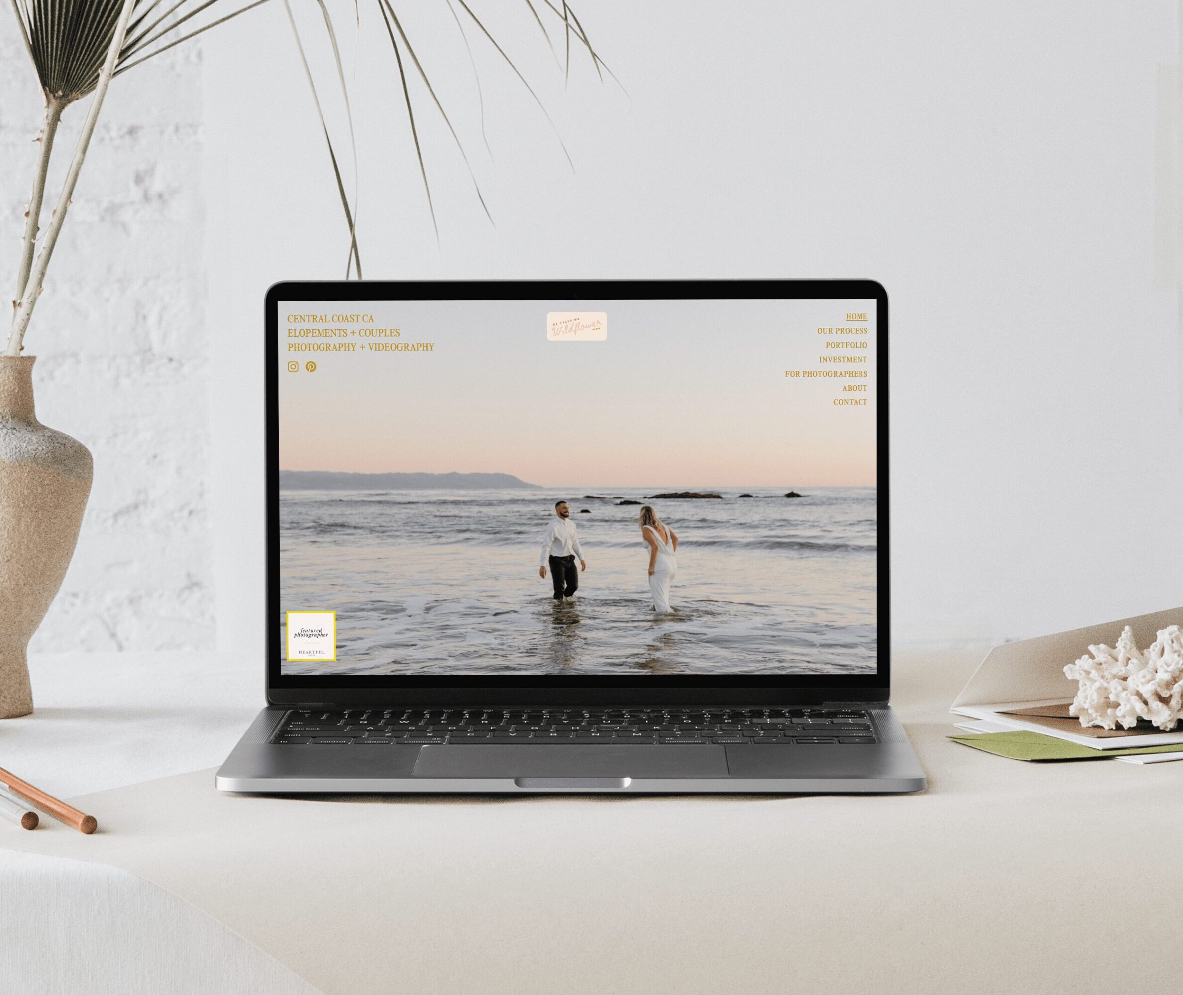 Laptop on a white desk displaying a beach scene with two people standing in the water, with a plant and a book nearby.