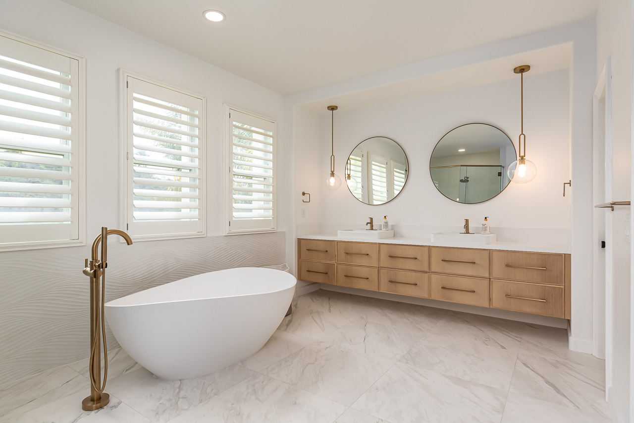 Bathroom with a freestanding bathtub, three windows with shutters, and a double vanity with two round mirrors, pendant lights, and wooden cabinets.