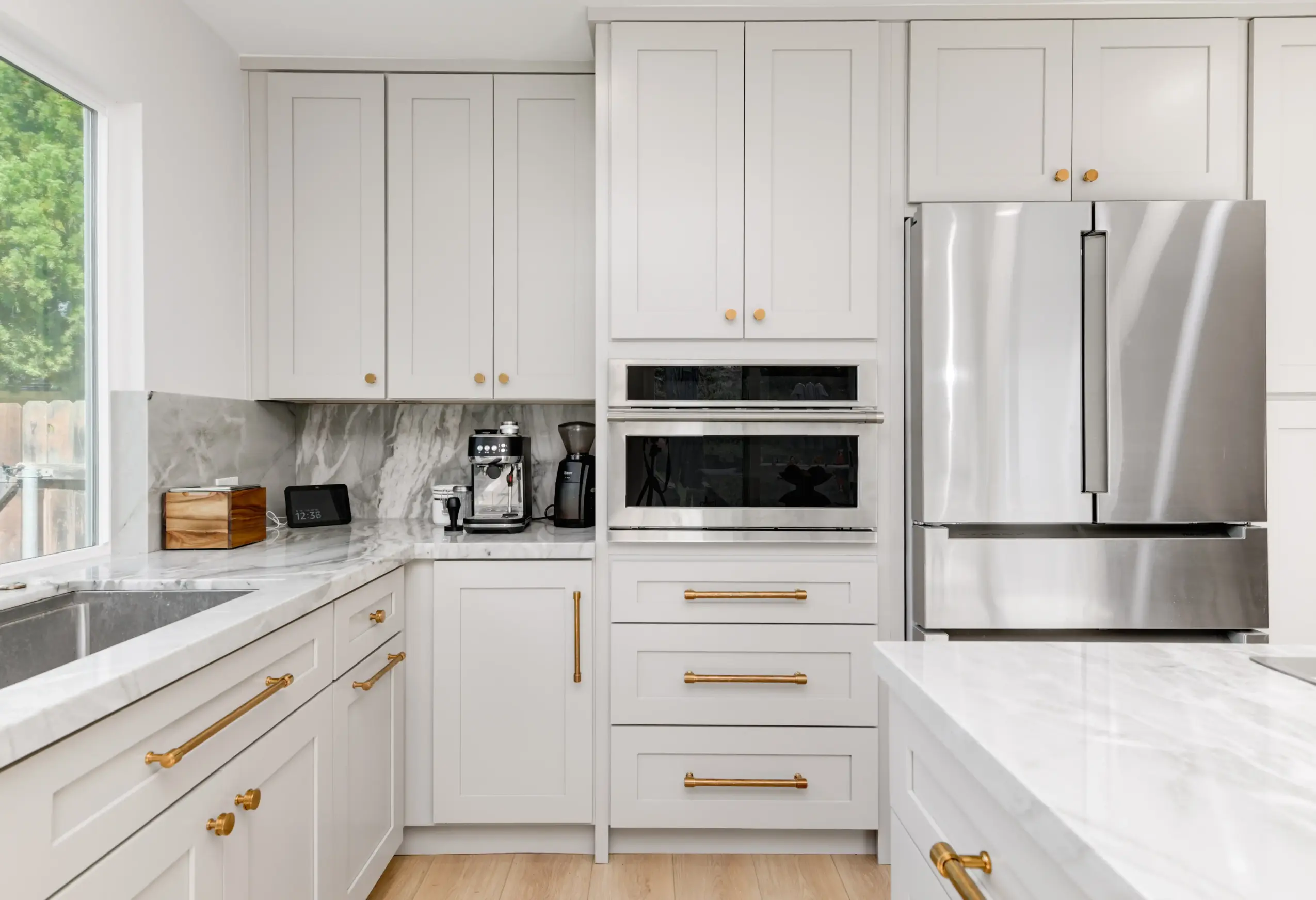 Modern kitchen with white cabinets, stainless steel refrigerator, and marble countertops, illuminated by natural light from a window.