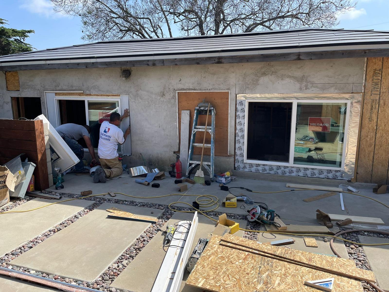 Construction workers installing a window on a building with unfinished exterior walls, tools and materials scattered on the ground.