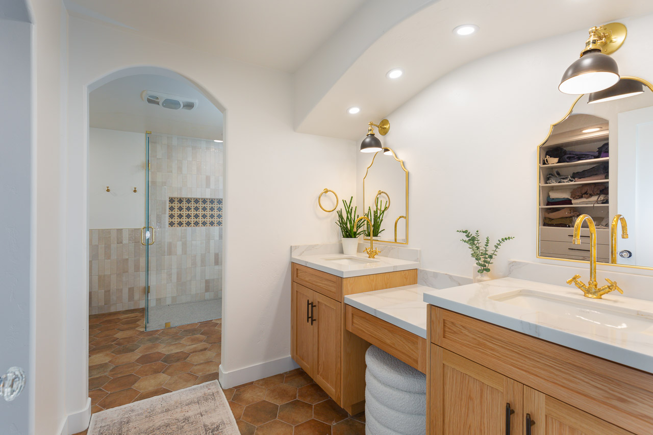 Bathroom vanity with two sinks, gold fixtures, wooden cabinets, and plants, with a shower area visible in the background.