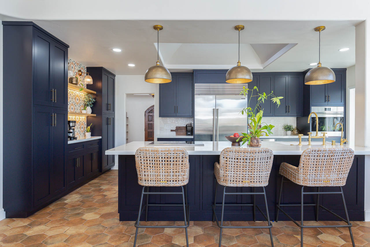 Modern kitchen with dark cabinets, white countertops, three bar stools, and pendant lights, with plants and appliances visible.