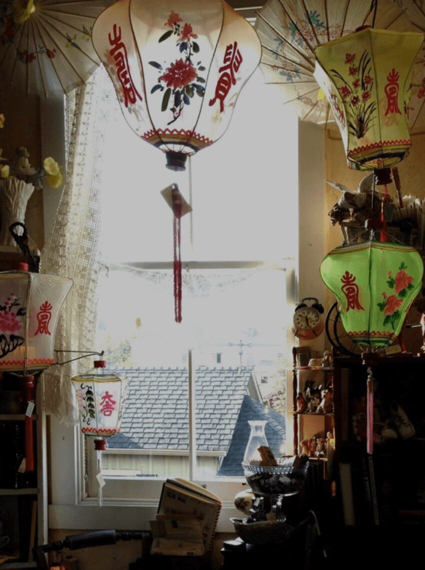 Indoor scene with hanging paper lanterns, window with curtains, and a view of rooftops outside, illuminated by natural light.