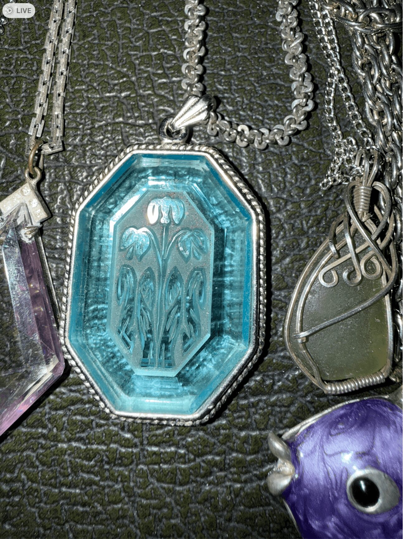 Pendant with blue glass and intricate design, surrounded by various silver chains and jewelry, on textured black surface.
