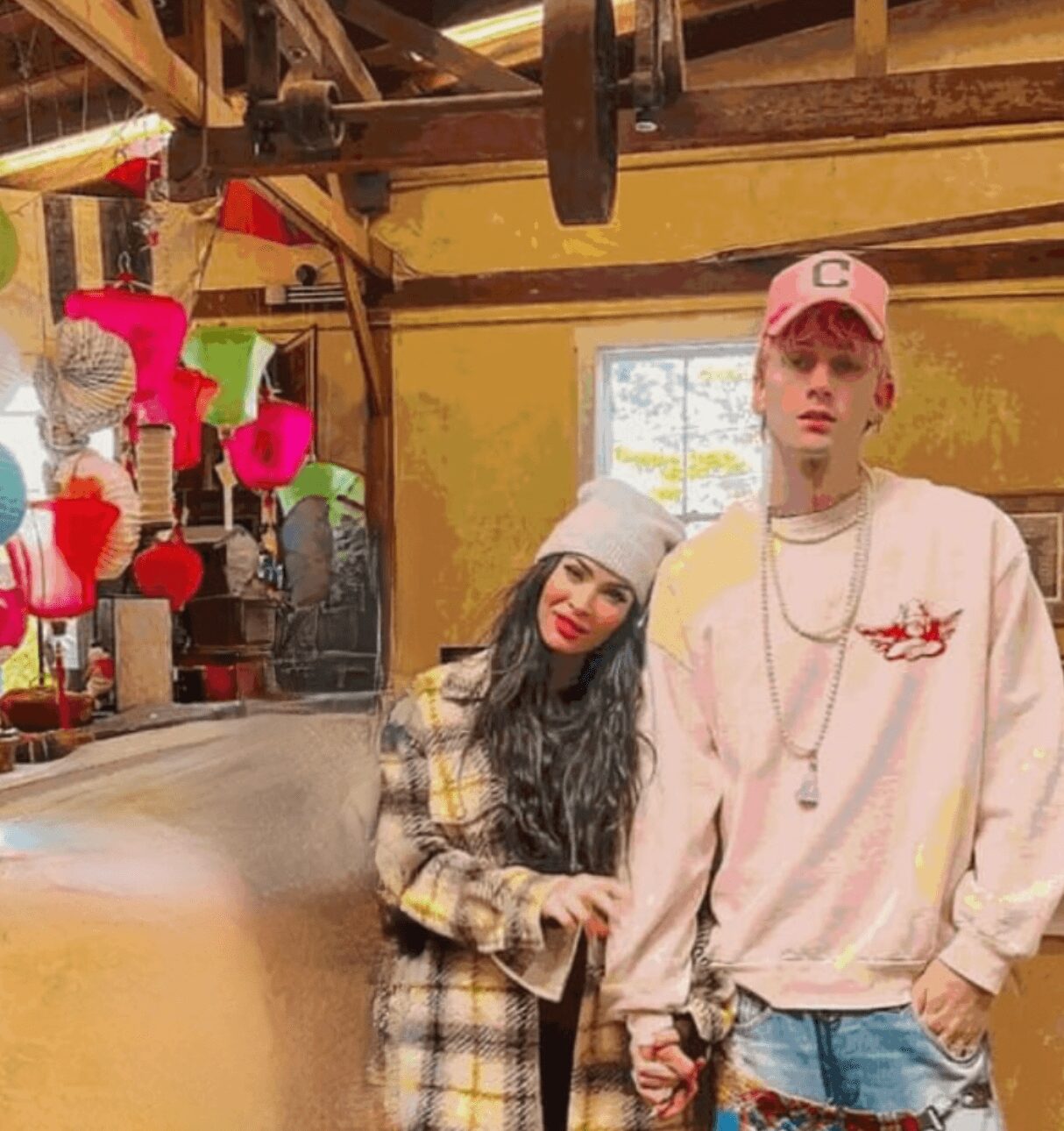 Two young people holding hands inside a rustic room with colorful decorations, one wearing a beanie and the other a pink cap.