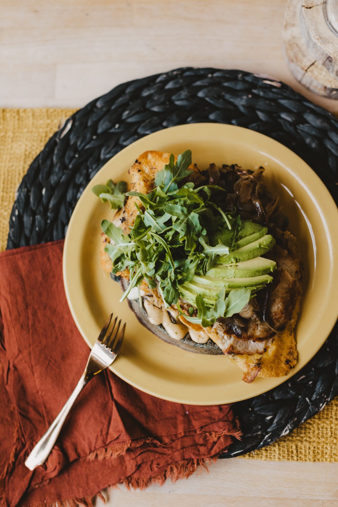 Plate with fish, greens, and vegetables on a yellow plate, on a black woven placemat, with a fork and cloth napkin.