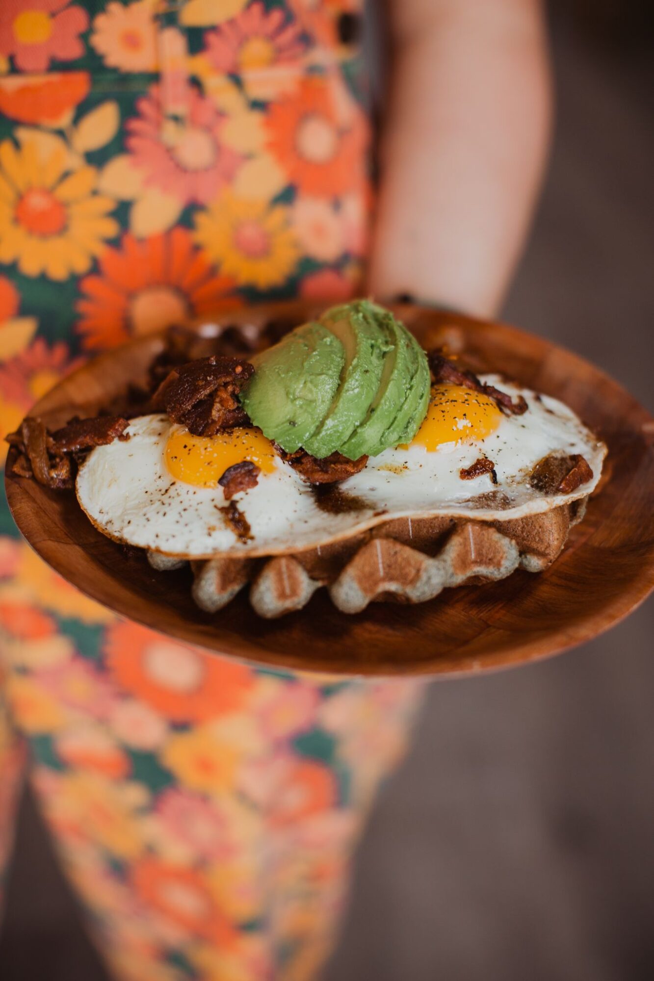 Person holding a wooden plate with a dessert topped with green ice cream, fruit, and chocolate, wearing a floral dress.