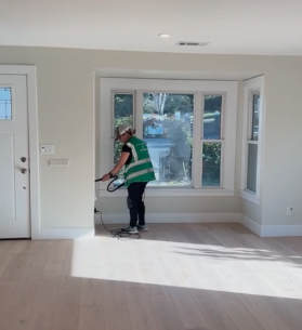 Person cleaning floor near large window in bright room with hardwood flooring, white walls, and a door on the left.