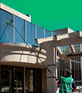 Person cleaning building windows with a long pole, wearing a green vest and black hat, against a building with glass and concrete.