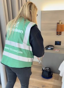 Person with blonde hair wearing a green vest labeled 'Clean' and 'Technician' standing in a room with a desk and bag.
