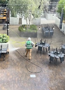 Person in green vest and khaki pants cleaning outdoor patio with chairs and trees in background.