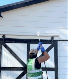 Person pressure washing the side of a building, wearing a cap, gloves, and a safety vest.